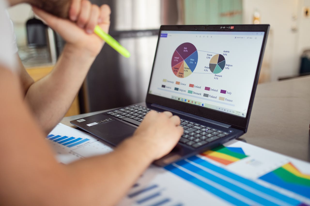 services-03 Person analyzing financial charts and graphs on a laptop with colorful documents, showcasing market analysis.